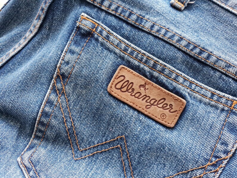May include: Close-up of blue denim jeans with a brown leather Wrangler patch on the back pocket. The patch features the Wrangler logo in a cursive font. The jeans have orange stitching.
