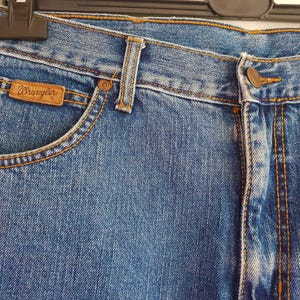 May include: Close-up of a pair of blue denim jeans with a brown leather Wrangler label. The jeans have a classic five-pocket style and a straight leg.