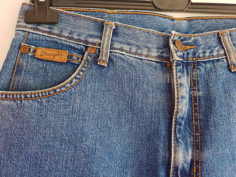 May include: Close-up of a pair of blue denim jeans with a brown leather Wrangler label. The jeans have a classic five-pocket style and a straight leg.