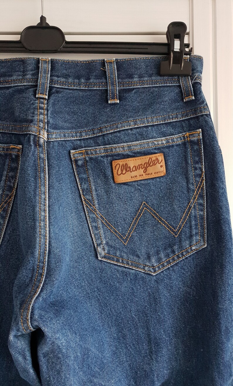 May include: A pair of blue denim jeans with a back pocket featuring a brown leather Wrangler label with the text "Wrangler Made for Tough Service".
