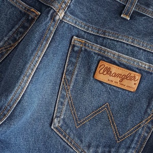 May include: Close-up of a blue denim back pocket with a brown leather patch that says "Wrangler Made for Tough Service."