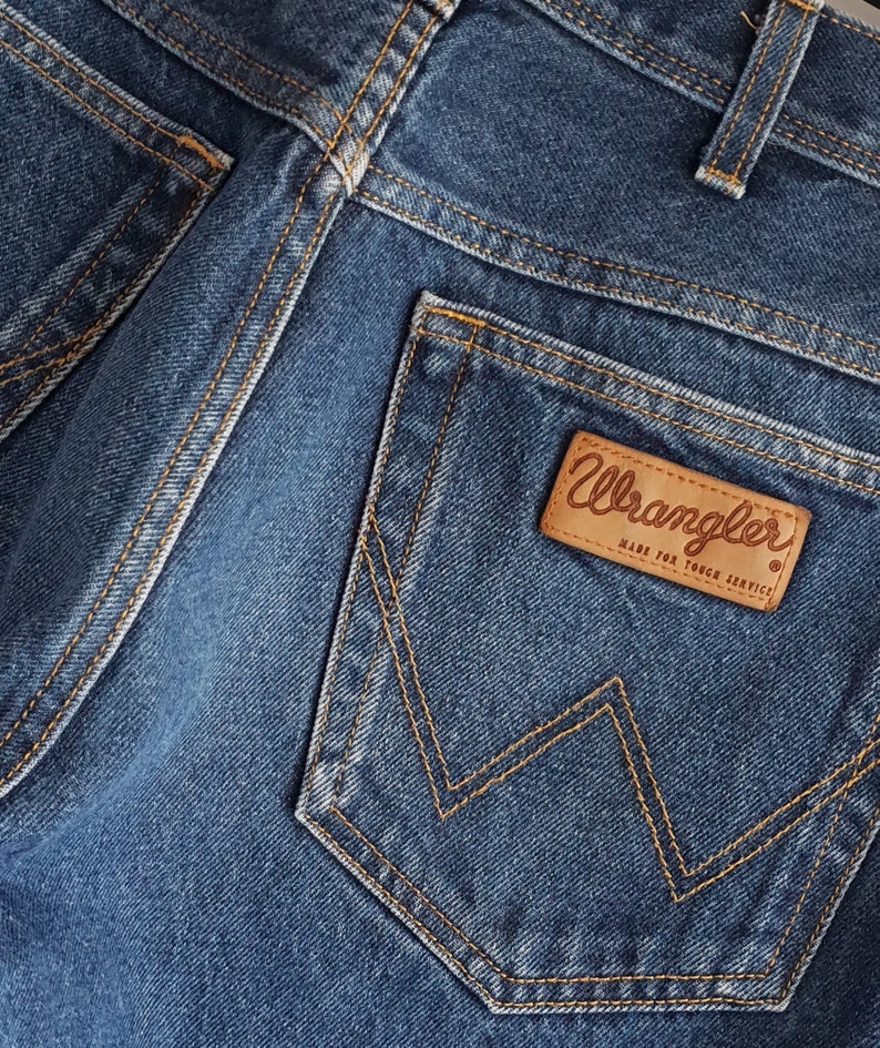 May include: Close-up of a blue denim back pocket with a brown leather patch that says "Wrangler Made for Tough Service."