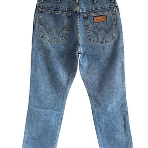 May include: Light blue denim jeans with the Wrangler logo on the back pocket. The jeans have a classic five-pocket design and a straight leg cut. The back pockets feature a signature stitching pattern.