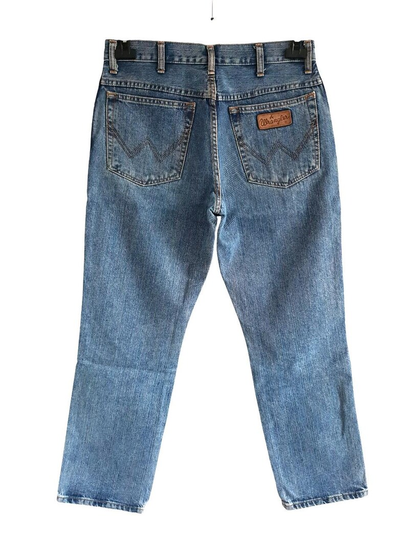 May include: Light blue denim jeans with the Wrangler logo on the back pocket. The jeans have a classic five-pocket design and a straight leg cut. The back pockets feature a signature stitching pattern.