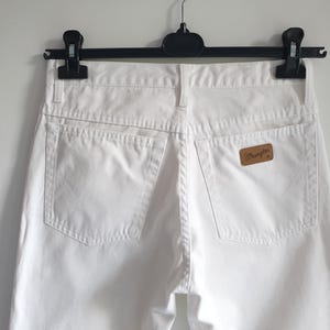 May include: Close-up of white denim jeans hanging on a black hanger. The jeans have two back pockets and a brown leather patch with the "Wrangler" logo. The jeans are made of a sturdy material and have a classic design.