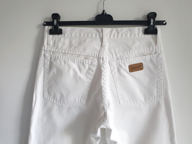 May include: Close-up of white denim jeans hanging on a black hanger. The jeans have two back pockets and a brown leather patch with the "Wrangler" logo. The jeans are made of a sturdy material and have a classic design.