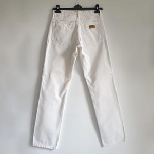 May include: White denim jeans with a classic five-pocket design. The jeans are hanging on a black hanger against a white background. A brown leather patch is visible on the back waistband.