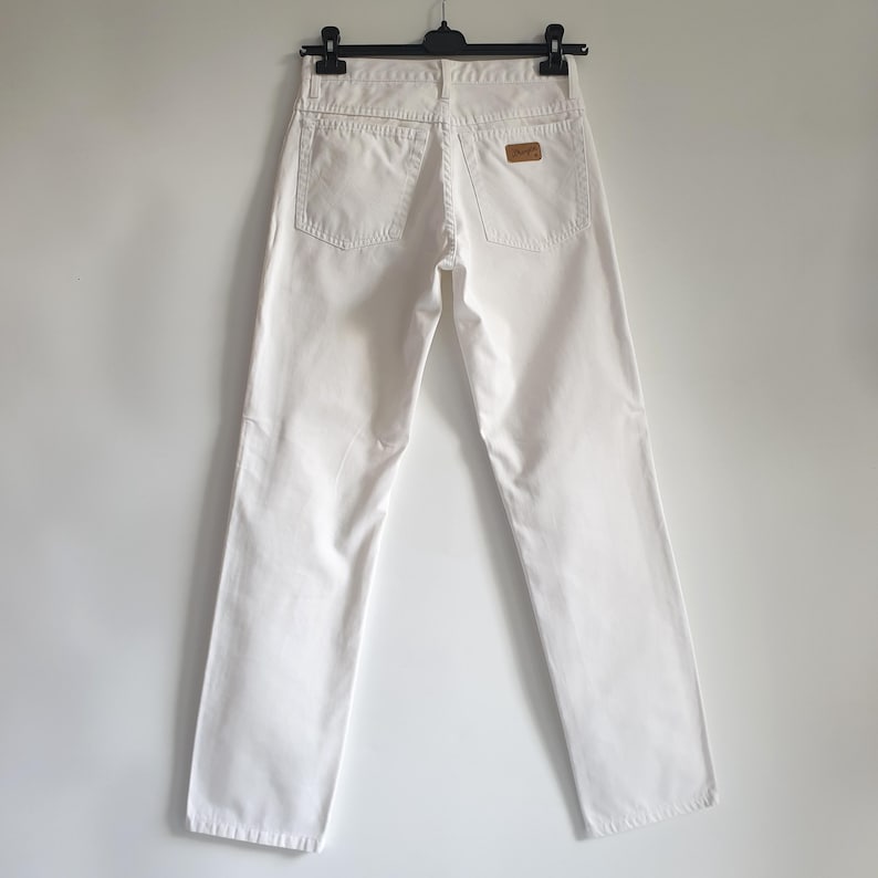 May include: White denim jeans with a classic five-pocket design. The jeans are hanging on a black hanger against a white background. A brown leather patch is visible on the back waistband.