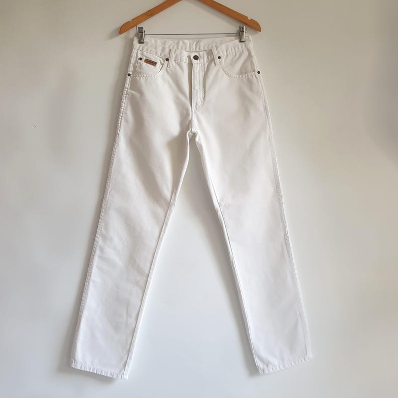 May include: White denim jeans hanging on a wooden hanger. The jeans have a classic five-pocket design with a button and zipper closure. The jeans are a solid white color and appear to be made of a sturdy denim material.