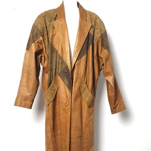 Può includere: Un cappotto vintage in pelle nei toni del marrone, beige e marrone scuro. Il cappotto presenta una silhouette lunga, un colletto a revers e una chiusura con un solo bottone. Le spalle e le tasche hanno un design a fantasia. Il cappotto è esposto su sfondo bianco.