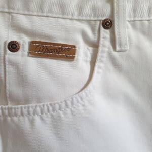 May include: Close-up of white denim jeans featuring a pocket with a brown leather tag that reads "Wrangler." The jeans have copper-colored rivets and stitching details. The fabric appears to be a sturdy cotton blend.