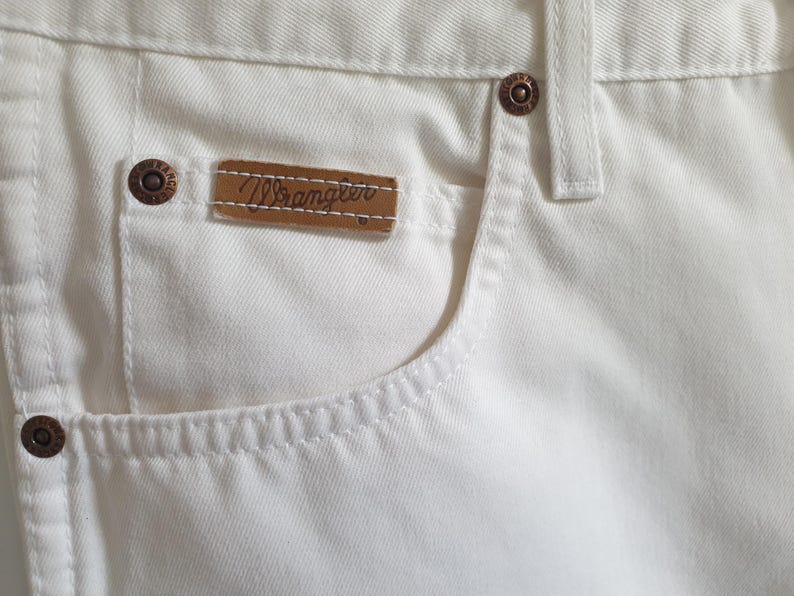 May include: Close-up of white denim jeans featuring a pocket with a brown leather tag that reads "Wrangler." The jeans have copper-colored rivets and stitching details. The fabric appears to be a sturdy cotton blend.