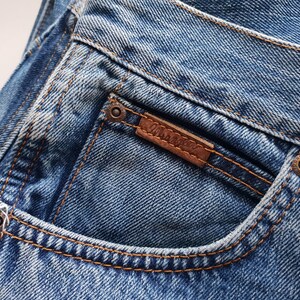 May include: Close-up of blue denim jeans with a brown leather patch and the brand name "Wrangler" stitched on the back pocket. The jeans have copper-colored rivets and stitching.