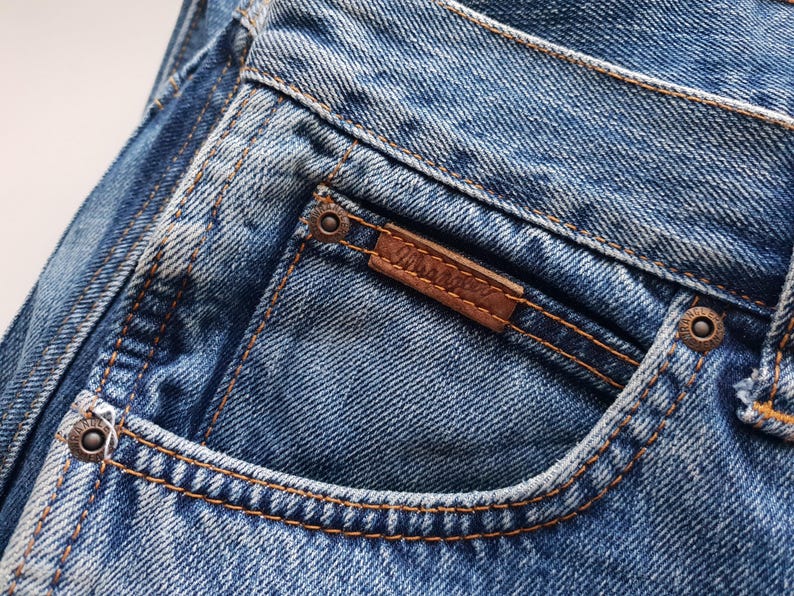 May include: Close-up of blue denim jeans with a brown leather patch and the brand name "Wrangler" stitched on the back pocket. The jeans have copper-colored rivets and stitching.