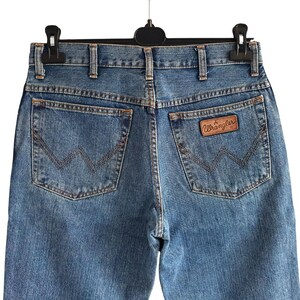 May include: Light blue denim jeans with the Wrangler logo on the back pocket. The jeans have classic five-pocket styling and a straight leg fit. The jeans are hanging on a black hanger.