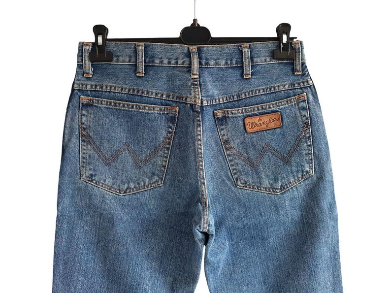 May include: Light blue denim jeans with the Wrangler logo on the back pocket. The jeans have classic five-pocket styling and a straight leg fit. The jeans are hanging on a black hanger.
