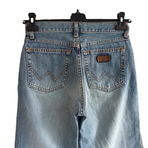 May include: Light blue denim jeans with classic five-pocket styling. The jeans feature a brown leather patch and the Wrangler logo on the back pocket. The jeans are hanging on a black hanger.