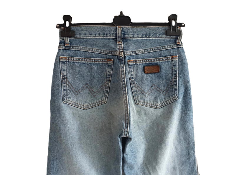 May include: Light blue denim jeans with classic five-pocket styling. The jeans feature a brown leather patch and the Wrangler logo on the back pocket. The jeans are hanging on a black hanger.