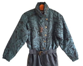 Könnte beinhalten: Ein blauer Ski-Overall mit einem abstrakten Schwarz-Weiß-Druck. Der Overall hat einen Reißverschluss, einen hohen Kragen und einen Gürtel.