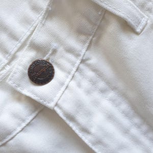 May include: Close-up of white denim fabric with a button closure. The button is a dark bronze color and has the word "Wrangler" embossed on it. The stitching is visible, and the fabric appears to be new.