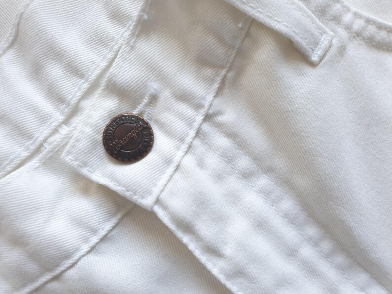 May include: Close-up of white denim fabric with a button closure. The button is a dark bronze color and has the word "Wrangler" embossed on it. The stitching is visible, and the fabric appears to be new.