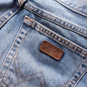 May include: Close-up of light blue denim jeans with a back pocket and a brown leather patch. The jeans have visible stitching in a contrasting orange-brown thread. The denim shows signs of wear and fading.