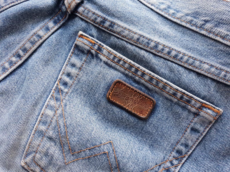 May include: Close-up of light blue denim jeans with a back pocket and a brown leather patch. The jeans have visible stitching in a contrasting orange-brown thread. The denim shows signs of wear and fading.