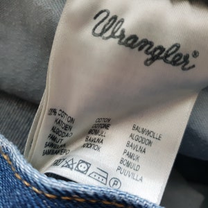 May include: A white fabric label with the word "Wrangler" printed in black. The label lists the word "cotton" in multiple languages, including English, French, German, Dutch, Spanish, Italian, Polish, and Portuguese.