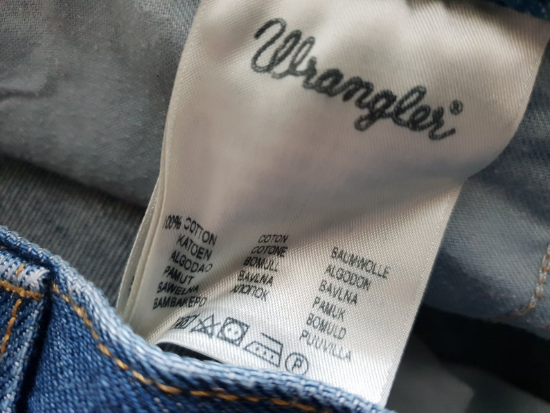 May include: A white fabric label with the word "Wrangler" printed in black. The label lists the word "cotton" in multiple languages, including English, French, German, Dutch, Spanish, Italian, Polish, and Portuguese.