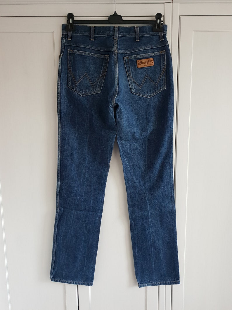 May include: A pair of blue denim jeans with a brown leather Wrangler label on the back pocket.