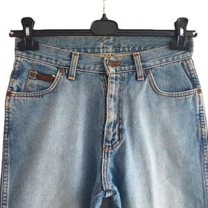 May include: Light blue denim jeans hanging on a black hanger. The jeans have a classic five-pocket design with a button and zipper closure. The jeans have a faded wash and a small brown leather patch on the back pocket.