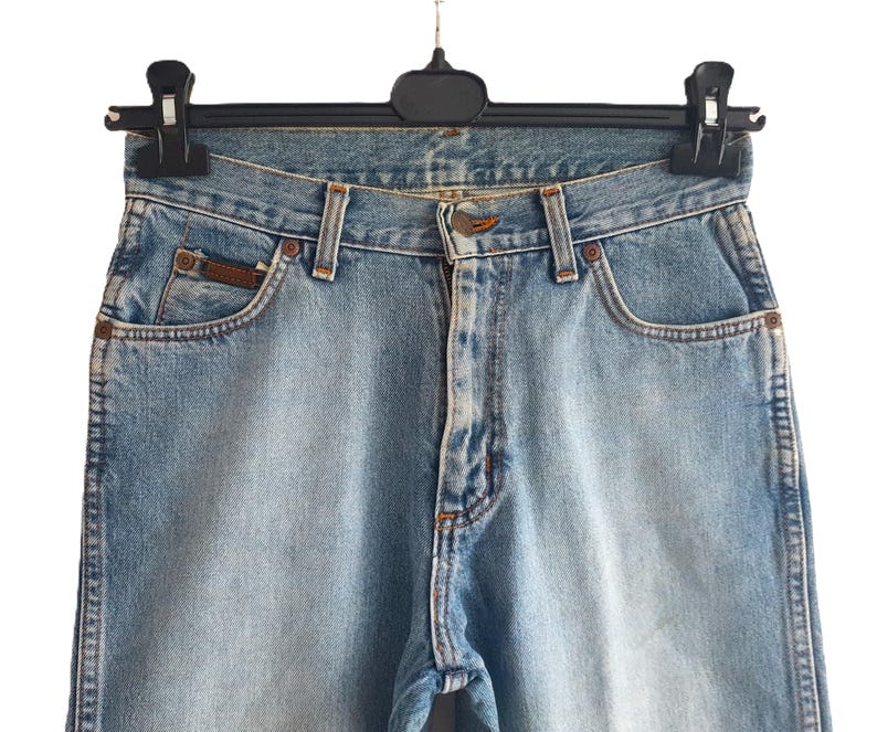 May include: Light blue denim jeans hanging on a black hanger. The jeans have a classic five-pocket design with a button and zipper closure. The jeans have a faded wash and a small brown leather patch on the back pocket.