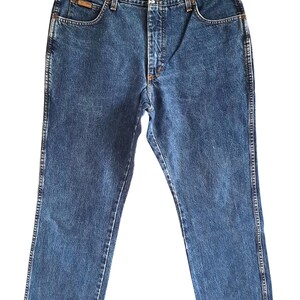 Op de afbeelding: Blauwe denim jeans met een klassieke rechte pijp. Kenmerken zijn een knoop- en ritssluiting, voor- en achterzakken en een kleine logopatch op de voorzak. De jeans hangen aan een zwarte hanger.