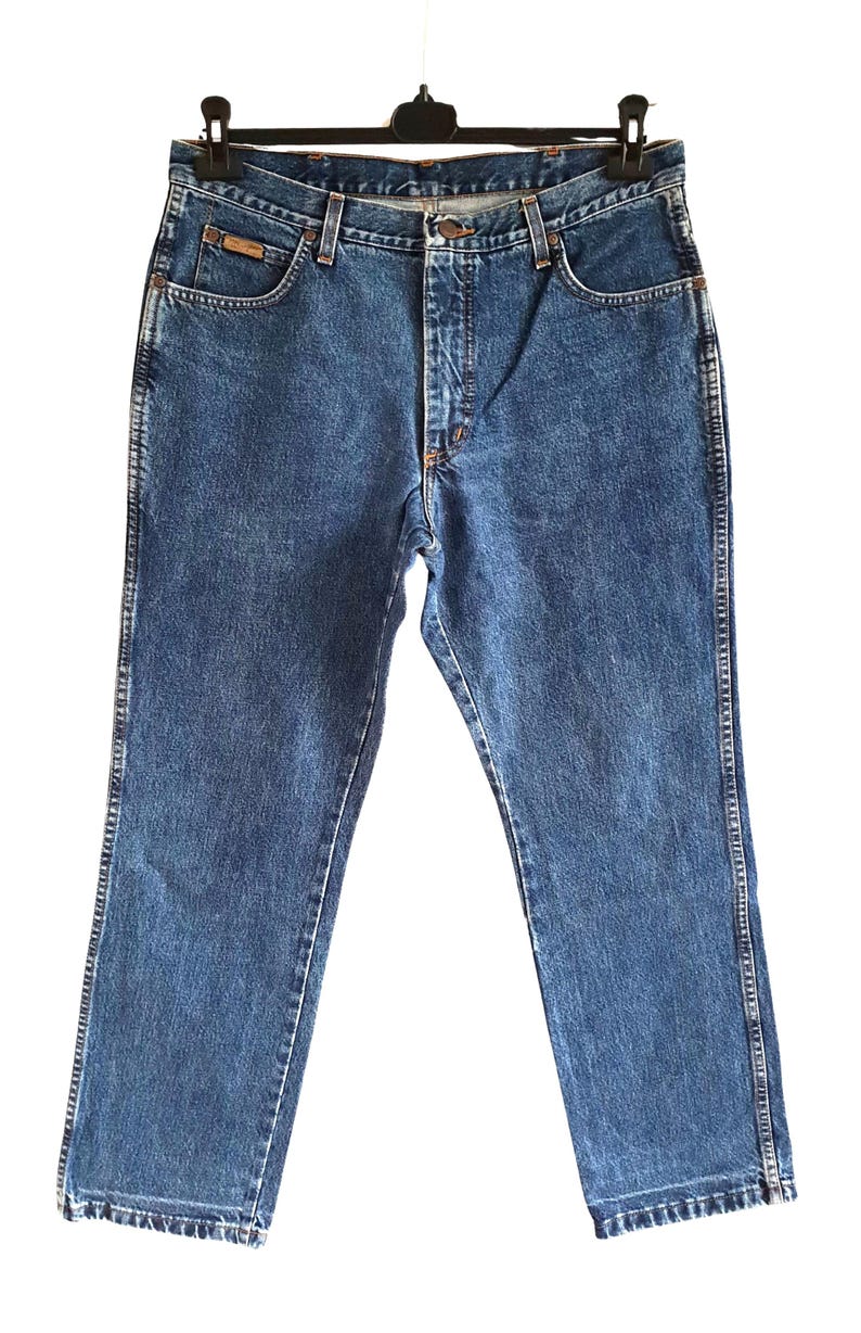Op de afbeelding: Blauwe denim jeans met een klassieke rechte pijp. Kenmerken zijn een knoop- en ritssluiting, voor- en achterzakken en een kleine logopatch op de voorzak. De jeans hangen aan een zwarte hanger.