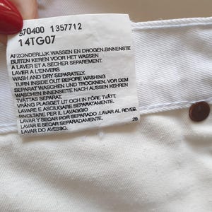 May include: Close-up of a white garment tag with care instructions in multiple languages. The tag includes the numbers "570400 1357712" and "14TG07". A brown button is visible on the garment. The tag also includes the number "20".