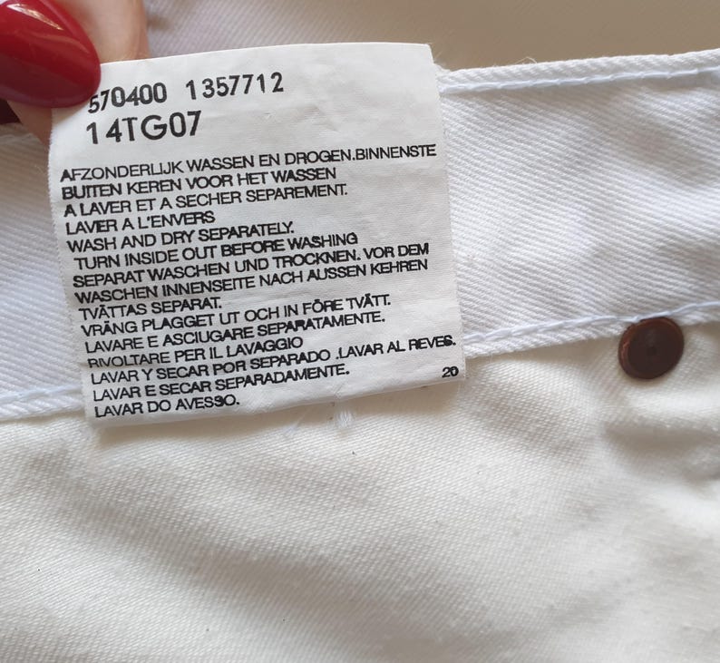 May include: Close-up of a white garment tag with care instructions in multiple languages. The tag includes the numbers "570400 1357712" and "14TG07". A brown button is visible on the garment. The tag also includes the number "20".