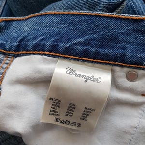 May include: Close-up of blue denim jeans with the brand label "Wrangler" visible. The label lists the fabric composition as 100% cotton in multiple languages. The jeans have orange stitching.