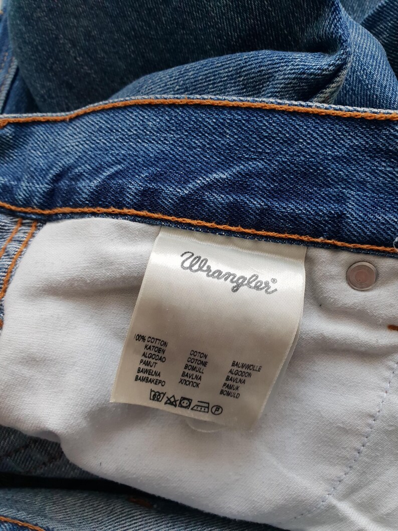 May include: Close-up of blue denim jeans with the brand label "Wrangler" visible. The label lists the fabric composition as 100% cotton in multiple languages. The jeans have orange stitching.
