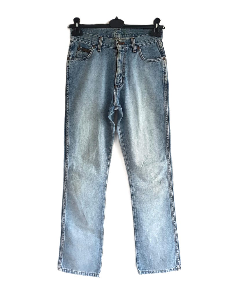 May include: Light blue denim jeans hanging on a black hanger. The jeans have a classic five-pocket design, a button and zipper closure, and a faded wash. The jeans are straight-legged and appear to be in good condition.