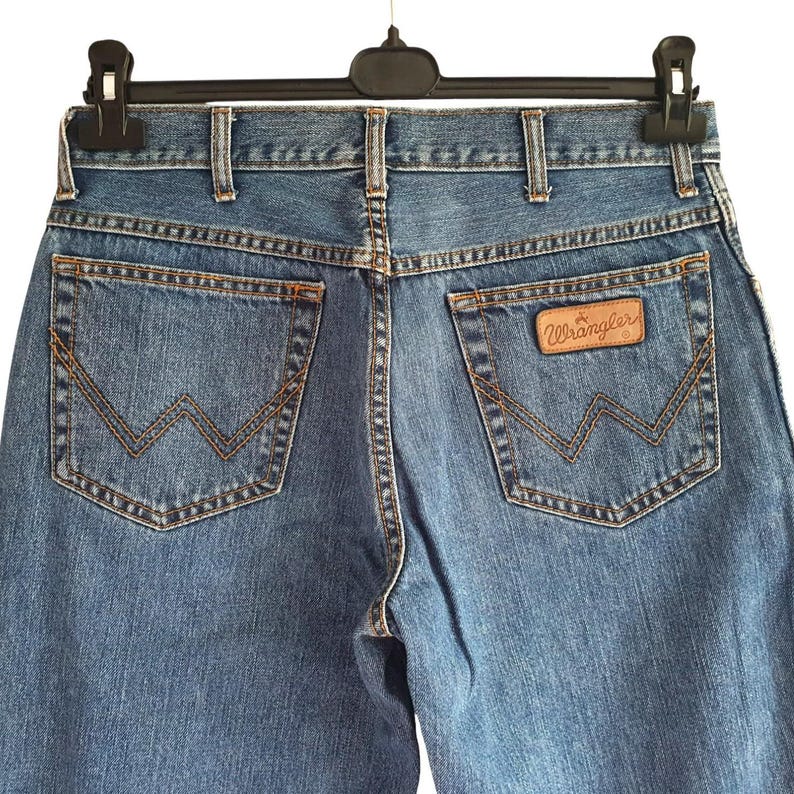 May include: A pair of blue denim jeans with a brown leather Wrangler label on the back pocket.