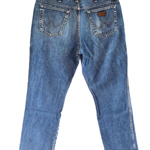 Puede incluir: Unos vaqueros de mezclilla en tono azul medio con cintura alta, trabillas para cintur&oacute;n y dos bolsillos traseros con una costura en forma de W. Un peque&ntilde;o parche de cuero marr&oacute;n es visible en el bolsillo trasero derecho, el pantal&oacute;n cuelga.