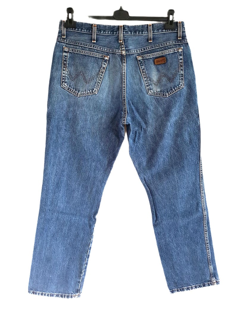 Puede incluir: Unos vaqueros de mezclilla en tono azul medio con cintura alta, trabillas para cintur&oacute;n y dos bolsillos traseros con una costura en forma de W. Un peque&ntilde;o parche de cuero marr&oacute;n es visible en el bolsillo trasero derecho, el pantal&oacute;n cuelga.