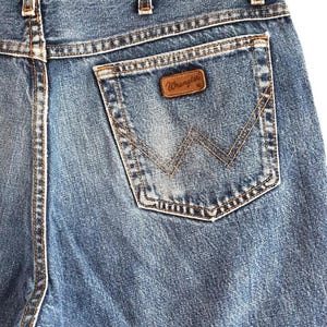 Puede incluir: Vaqueros de mezclilla azul con dise&ntilde;o de cintura alta, trabillas para cintur&oacute;n y un bolsillo trasero. El bolsillo muestra un parche de cuero marr&oacute;n con el texto Wrangler y un patr&oacute;n de costura decorativo en forma de W en hilo color cobre.