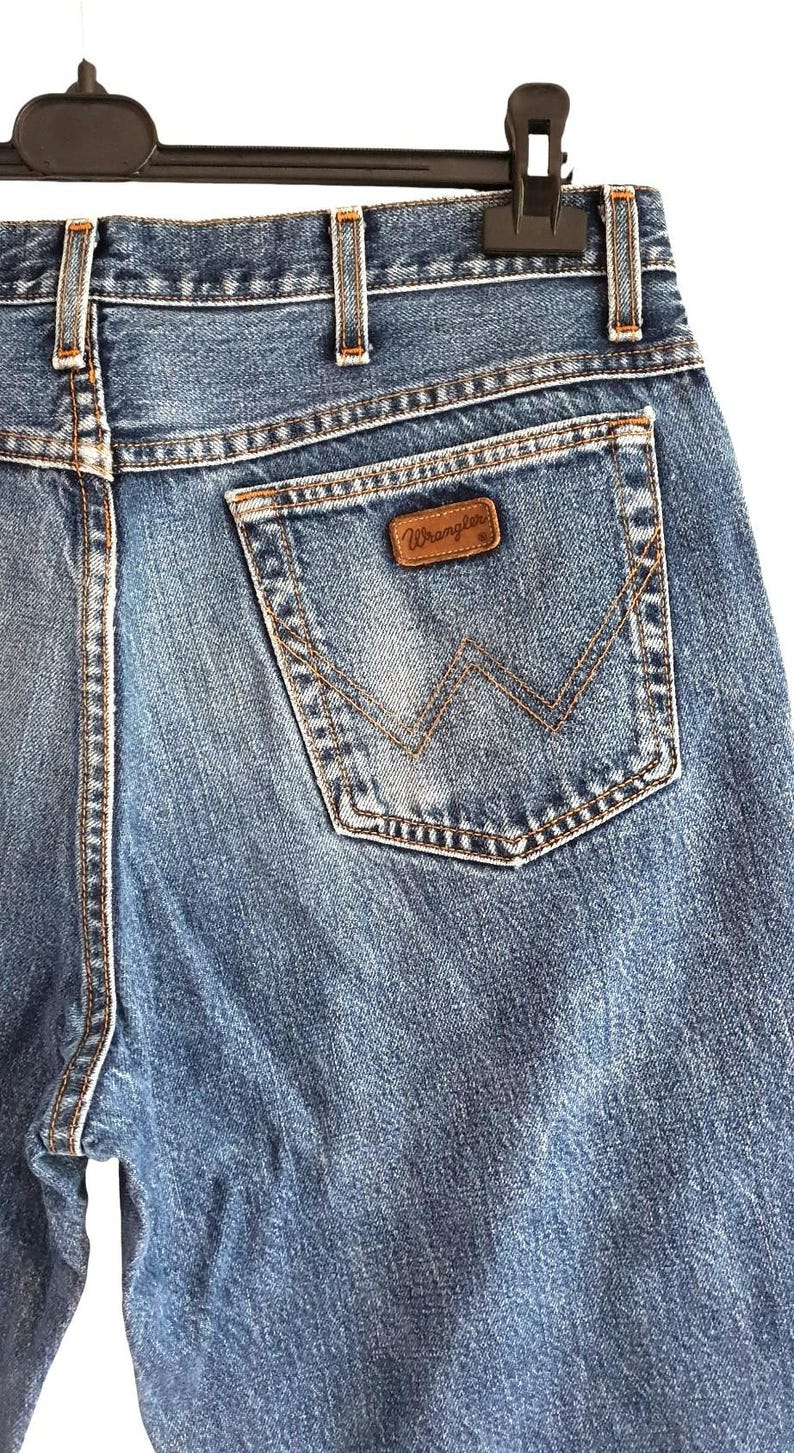 Puede incluir: Vaqueros de mezclilla azul con dise&ntilde;o de cintura alta, trabillas para cintur&oacute;n y un bolsillo trasero. El bolsillo muestra un parche de cuero marr&oacute;n con el texto Wrangler y un patr&oacute;n de costura decorativo en forma de W en hilo color cobre.