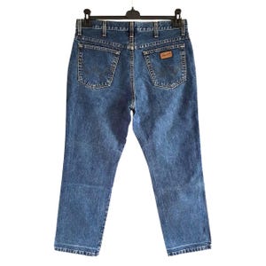 Op de afbeelding: Blauwe spijkerbroek hangt aan een zwarte hanger. De jeans heeft een klassiek ontwerp met vijf zakken, met de Wrangler-logopatch op de achterzak. De jeans heeft een medium wassing en lijkt in goede staat te verkeren.
