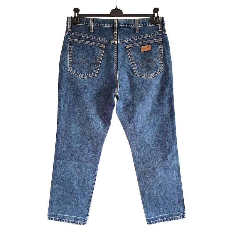 Op de afbeelding: Blauwe spijkerbroek hangt aan een zwarte hanger. De jeans heeft een klassiek ontwerp met vijf zakken, met de Wrangler-logopatch op de achterzak. De jeans heeft een medium wassing en lijkt in goede staat te verkeren.