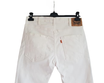 May include: Off-white denim jeans hanging on a black hanger. The jeans have two back pockets and a small red tag on the right back pocket. A brown leather patch with a logo is visible on the back waistband.