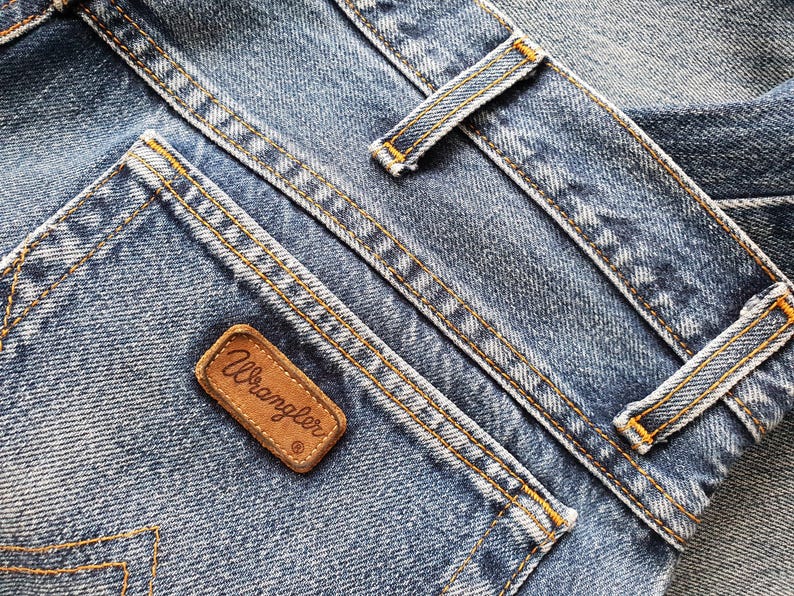 Puede incluir: Vaqueros de mezclilla azul con un lavado medio, textura visible y costuras en contraste de color naranja. Un parche rectangular de cuero marr&oacute;n con la palabra Wrangler grabada en letra cursiva est&aacute; cosido en el bolsillo trasero, junto a trabillas.