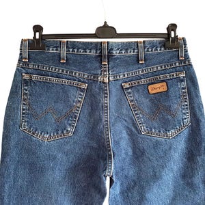 Op de afbeelding: Close-up van de achterkant van een blauwe spijkerbroek die aan een zwarte hanger hangt. De jeans heeft twee achterzakken met een gestikt ontwerp en een bruin leren label met de merknaam "Wrangler".