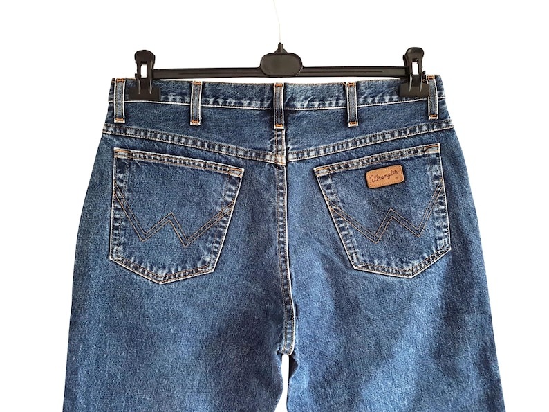 Op de afbeelding: Close-up van de achterkant van een blauwe spijkerbroek die aan een zwarte hanger hangt. De jeans heeft twee achterzakken met een gestikt ontwerp en een bruin leren label met de merknaam "Wrangler".